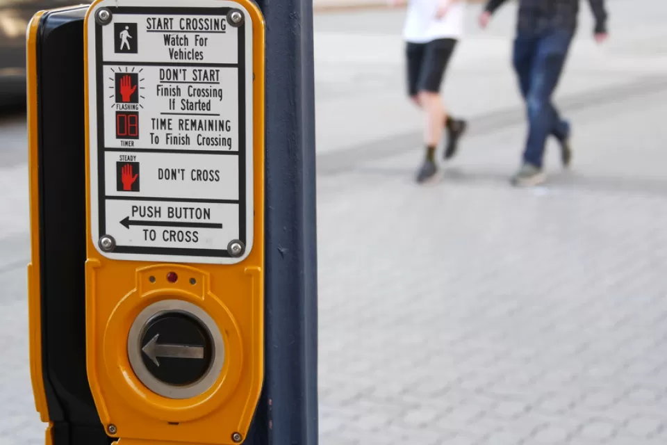 A crosswalk activation button is shown in the foreground with two pedestrians show blurred while crossing the street in the background.